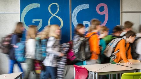 An einer Tafel in einem Klassenzimmer steht links "G8" und rechts "G9" geschrieben. Schülerinnen und Schüler laufen vor der Tafel auf die rechte Seite der Tafel, auf der "G9" steht.  (Foto: dpa Bildfunk, picture alliance / Armin Weigel/dpa | Armin Weigel)