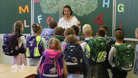 Lehrerin steht vor ihrer Klasse. Alle Kinder haben Schulranzen auf. (Foto: dpa Bildfunk, Picture Alliance)