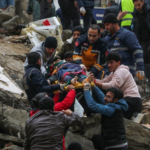 Menschen und Rettungskräfte bergen eine Person auf einer Bahre aus einem eingestürzten Gebäude. (Foto: dpa Bildfunk, Picture Alliance)