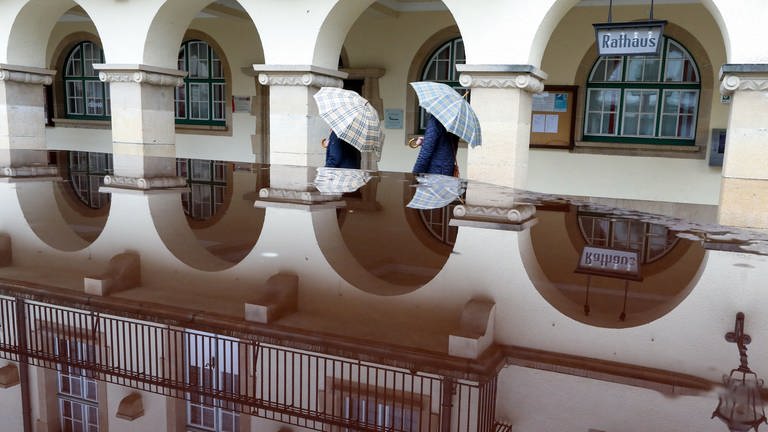 In einer Regenpfütze spiegelt sich das Rathaus von Sigmaringen wieder, an dem gerade zwei Passanten mit Regenschirmen vorbei gehen. (Foto: dpa Bildfunk, picture alliance/dpa | Thomas Warnack)