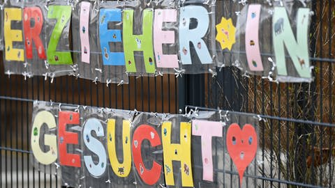Mit bunten Buchstaben sind die Worte "Erzieher*in gesucht" geformt, die an einem Zaun einer Kindertagesstätte befestigt sind.  (Foto: dpa Bildfunk, picture alliance/dpa | Arne Dedert)