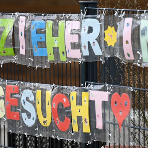 Mit bunten Buchstaben sind die Worte "Erzieher*in gesucht" geformt, die an einem Zaun einer Kindertagesstätte befestigt sind.  (Foto: dpa Bildfunk, picture alliance/dpa | Arne Dedert)