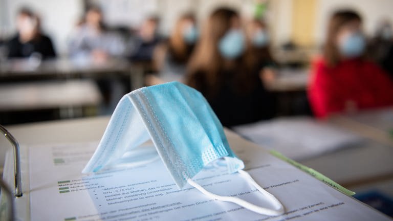 ine Maske liegt im Unterricht in einem Gymnasium auf Unterrichtsunterlagen, während im Hintergrund Schülerinnen und Schüler mit Mund- und Nasenschutz zu sehen sind. (Foto: dpa Bildfunk, picture alliance/Matthias Balk/dpa)