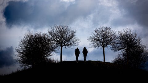 Spaziergänger gehen auf einen Hügel am Kronsberg, während bei stürmischem Wetter dunkle Wolken am Horizont vorüberziehen. (Foto: dpa Bildfunk, picture alliance/dpa | Moritz Frankenberg (Symbolfoto))
