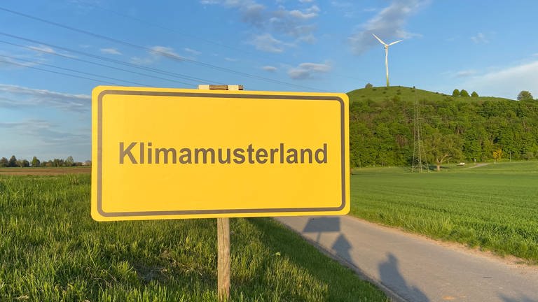 Ein gelbes Schild mit der schwarzen Aufschrift "Klimamusterland" steht vor einem Hügel mit einem Windrad darauf.  (Foto: SWR, Luisa Funk)