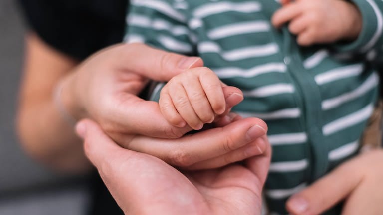Ein Mutter und ein Vater halten die Hand ihres neugeborenen Babys. (Foto: IMAGO, IMAGO / Cavan Images)