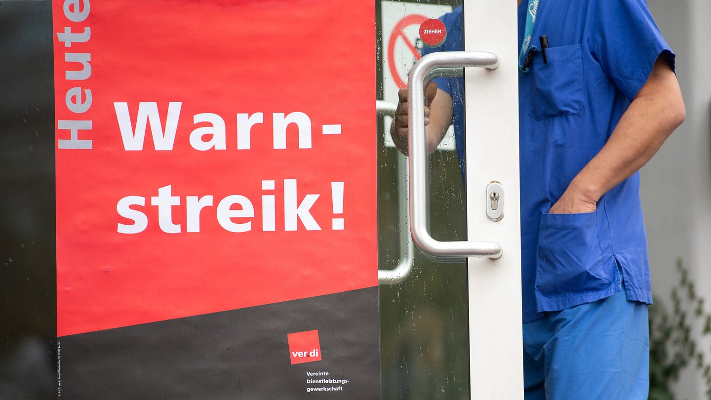 Ein Mitarbeiter des Universitätsklinikums Tübingen geht während eines Warnstreiks an den Uni-Kliniken durch eine Tür mit einem Plakat, auf dem 