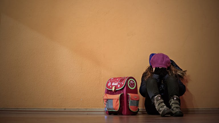 Ein Mädchen sitzt neben ihrem Schulranzen auf dem Boden und hält die Hände vor das Gesicht.