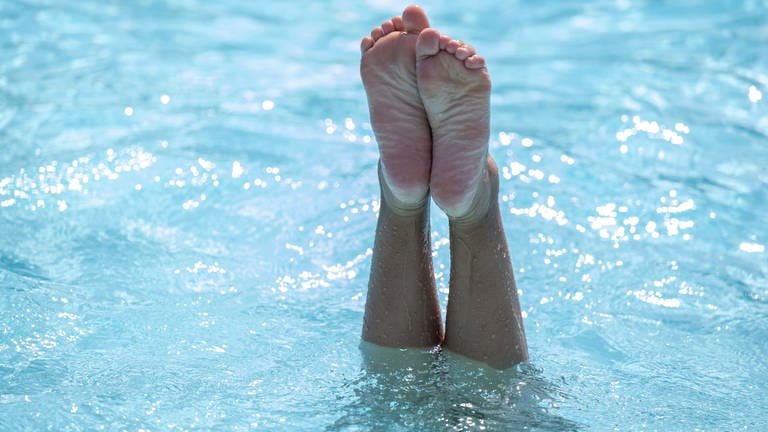 Eine Frau taucht in einem Schwimmbad kopfüber ins Wasser ein - über der Wasseroberfläche sind nur noch die Füße zu sehen.