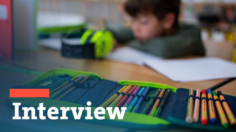Ein Mäppchen liegt aufgeschlagen mit vielen Stiften darin auf einem Schulpult, während ein Schüler im Hintergrund in sein Heft schreibt.