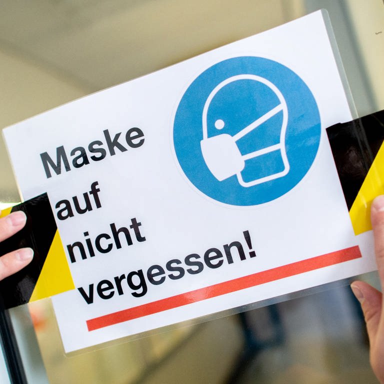Eine Person klebt ein Schild mit der Aufschrift "Maske auf nicht vergessen!" an die Glasscheibe einer Zwischentür. (Foto: dpa Bildfunk, picture alliance/dpa | Hauke-Christian Dittrich)