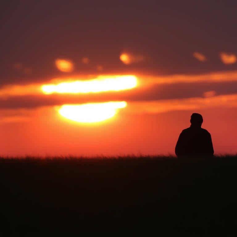 Silhouetten zweier Menschen vor dem Sonnenaufgang, der teilweise von Wolken bedeckt wird. (Foto: dpa Bildfunk, picture alliance/dpa | Thomas Warnack)