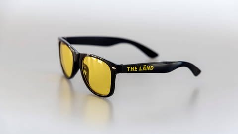 Eine Brille mit dem Werbeslogan "THE LÄND" liegt auf einem Tisch. (Foto: IMAGO, IMAGO / Arnulf Hettrich)