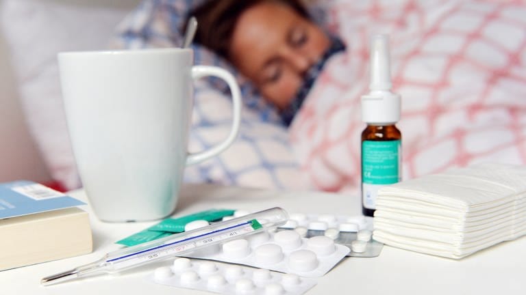 Eine Frau liegt mit Grippe im Bett, auf dem Nachtisch Fieberthermometer, Teetasse, Taschentücher, Tabletten und Nasenspray (Foto: dpa Bildfunk, Maurizio Gambarini)