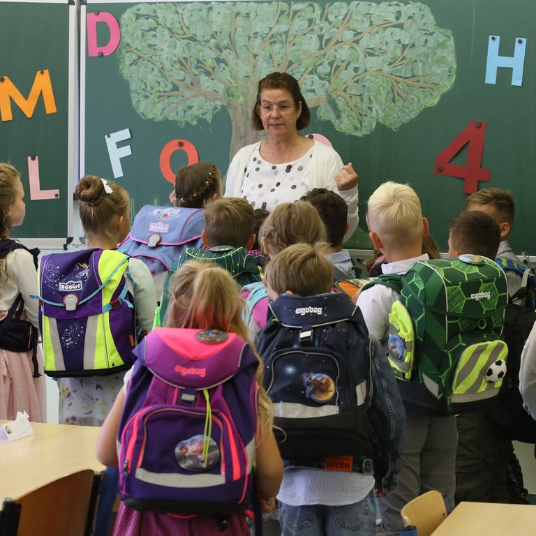Lehrerin steht vor ihrer Klasse. Alle Kinder haben Schulranzen auf. (Foto: dpa Bildfunk, Picture Alliance)