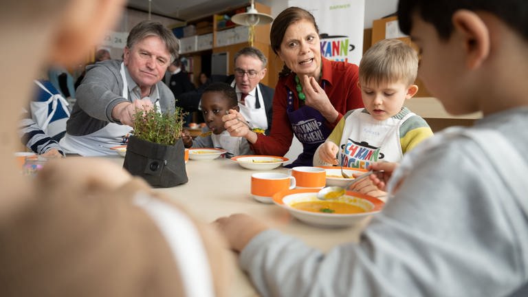 BW-Sozialminister Manne Lucha (Grüne), Christoph Strauch, Vorstandsvorsitzender der Barmer und Promi-Köchin Sarah Wiener sitzen mit Kindern an einem Tisch.