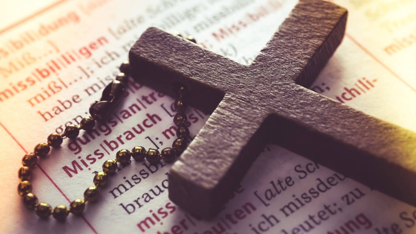Symbolbild für Missbrauch in der katholischen Kirche: Ein Kreuz liegt auf einem Wörterbuch (Eintrag Missbrauch) (Foto: IMAGO, IMAGO / Christian Ohde)