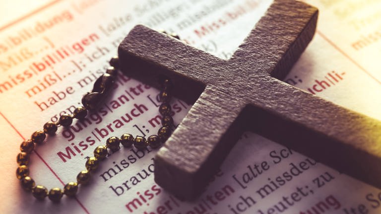 Symbolbild für Missbrauch in der katholischen Kirche: Ein Kreuz liegt auf einem Wörterbuch (Eintrag Missbrauch) 