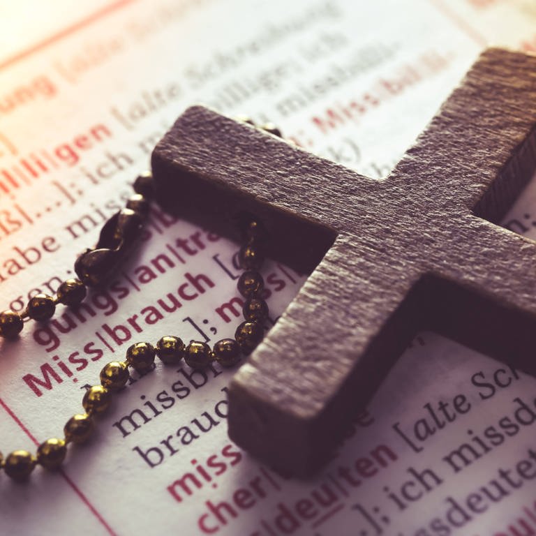 Symbolbild für Missbrauch in der katholischen Kirche: Ein Kreuz liegt auf einem Wörterbuch (Eintrag Missbrauch)  (Foto: IMAGO, IMAGO / Christian Ohde)