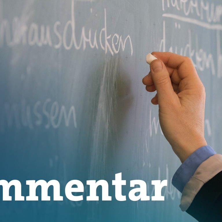 Eine Lehrerin steht in einem Klassenraum an einer Tafel und schreibt. Im Vordergrund steht: Kommentar.