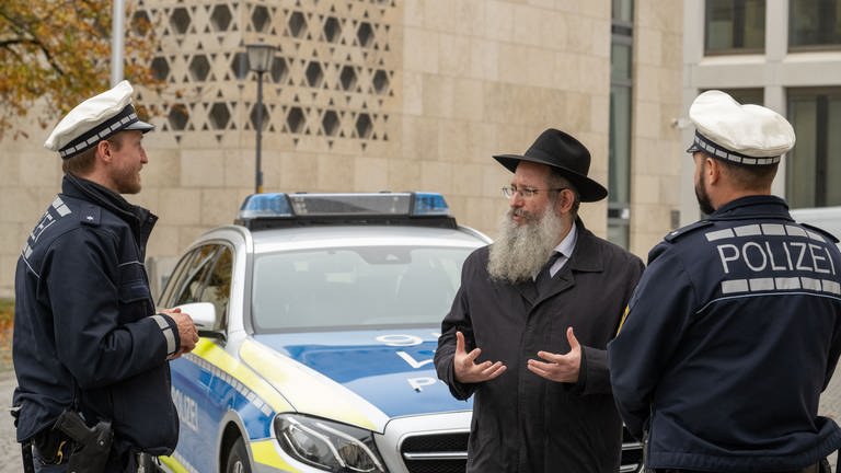 Rabbiner Shneur Trebnik steht mit zwei Polizisten vor der Synagoge an einem Polizeiauto. (Foto: dpa Bildfunk, picture alliance/dpa | Stefan Puchner)