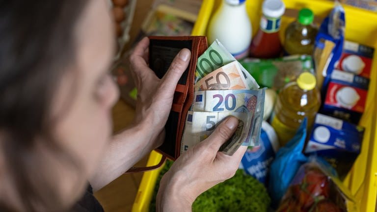 Eine Frau hält Geldscheine in einer Geldbörse über einer Einkaufskiste mit Lebensmitteln, der auf einem Küchentisch steht. (Foto: dpa Bildfunk, picture alliance/dpa | Hendrik Schmidt)