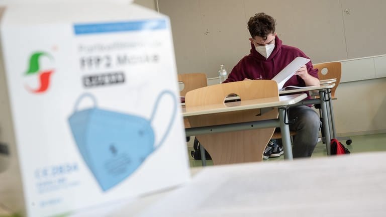 Ein Schüler mit medizinischer Maske liest sich in einem Klassenzimmer die Abituraufgaben durch.