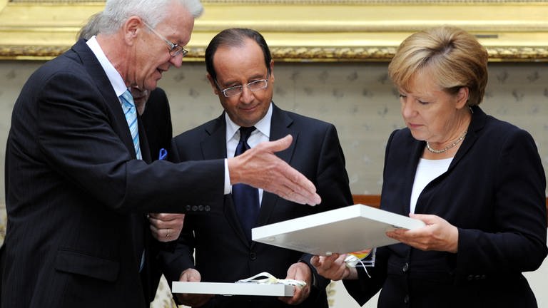 Winfried Kretschmann überreicht dem ehemaligen französische Staatspräsident Francois Hollande und der ehemaligen Bundeskanzlerin Angela Merkel (CDU) ein Gastgeschenk. (Foto: dpa Bildfunk, picture alliance / dpa | Bernd Weissbrod)