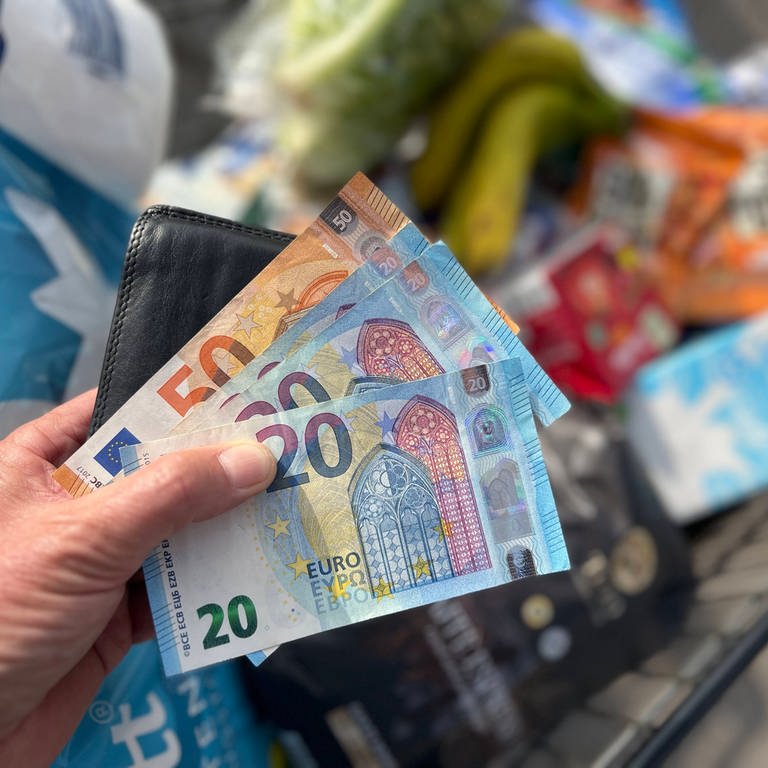 Geldscheine über einem Einkaufswagen der mit Lebensmitteln gefüllt ist. (Foto: IMAGO, IMAGO / Sven Simon)