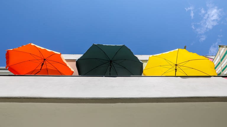 Drei Sonnenschirme stehen auf einem Balkon an einem Mehrfamilienhaus.