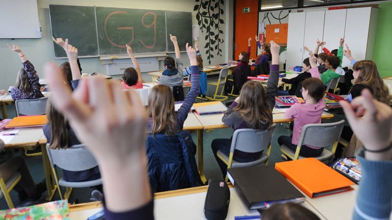 Schülerinnen und Schüler sitzen in einem Unterrichtsraum und heben ihre Finger, an der Tafel steht "G9". 