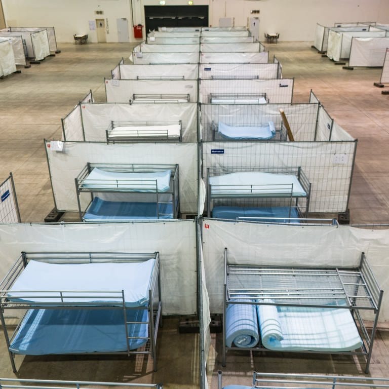 Stockbetten stehen zwischen Zäunen in einer Notunterkunft für Asylsuchende: Städte und Gemeinden in BW suchen händeringend nach Unterkünften für Geflüchtete (Symbolbild).