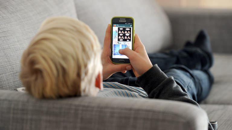 Ein Jugendlicher liegt auf einem Sofa und schaut auf sein Smartphone.
