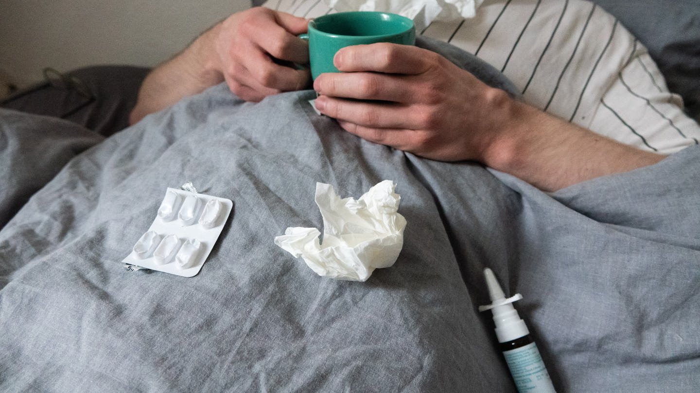 Ein Mann liegt mit Taschentüchern, Teetasse, Nasenspray und Tabletten im Bett. (Foto: dpa Bildfunk, picture alliance/dpa | Philip Dulian)