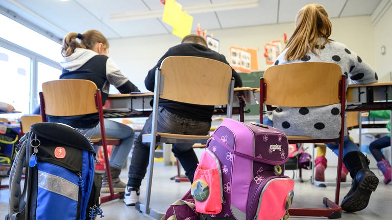 Schulranzen stehen hinter den Stühlen von drei Schülern in einer Grundschule in Stuttgart. (Foto: dpa Bildfunk, picture alliance/dpa | Sebastian Gollnow)