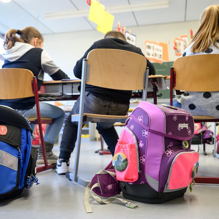 Schulranzen stehen hinter den Stühlen von drei Schülern in einer Grundschule in Stuttgart.