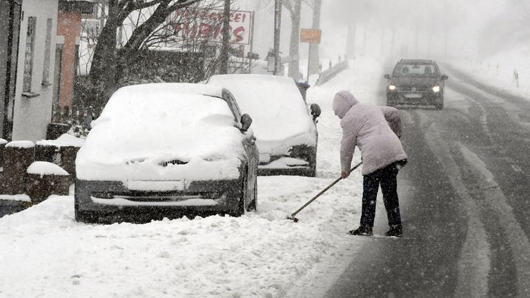 Eine Frau schippt auf einem Bürgersteig Schnee. Heute gibt der Winter noch mal ein kurzes Gastspiel, dabei fällt Schnee bis in Tieflagen.