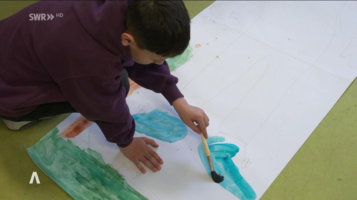 Ein Junge malt mit Wasserfarben auf dem Boden ein Selbstporträt aus. In der LEA in Ellwangen malen geflüchtete Kinder. Es ist zugleich Therapie und Beschäftigung. (Foto: SWR)