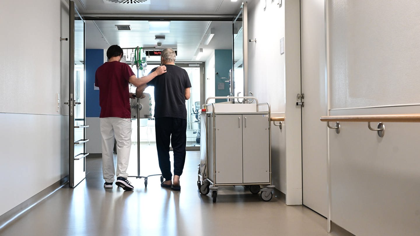 Ein Pfleger geht im Klinikum Stuttgart mit einem Patienten über einen Flur. (Foto: dpa Bildfunk, picture alliance/dpa | Bernd Weißbrod)