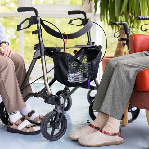 Symbolbild | Zwei Bewohnerinnen eines Altenheims sitzen neben ihren Rollatoren auf roten Sesseln. Ihre Füße stecken in Sandalen und Hausschuhen. 