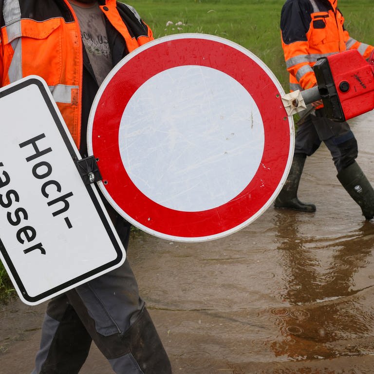 Mitarbeiter der Stadt Riedlingen stellen Absperr- und Hinweisschilder mit der Aufschrift "Hochwasser"an einem vom Regen überfluteten Radweg auf.