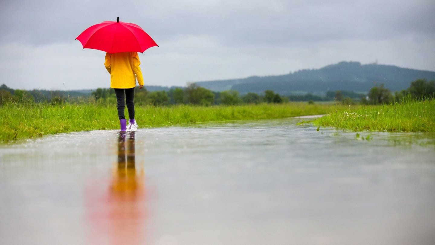Eine Spaziergängerin in Gummistiefeln läuft im Regen auf einem überfluteten Weg entlang. (Foto: dpa Bildfunk, picture alliance/dpa | Thomas Warnack)
