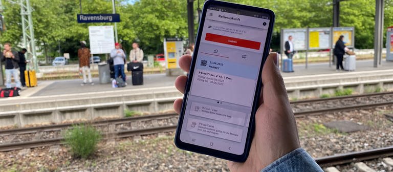 Seit Mittwoch gilt das 9-Euro-Ticket. Auf einem Handy ist die digitale Version zu sehen. Im Hintergrund der Bahnhof Ravensburg. (Foto: SWR, Thorben Langwald)