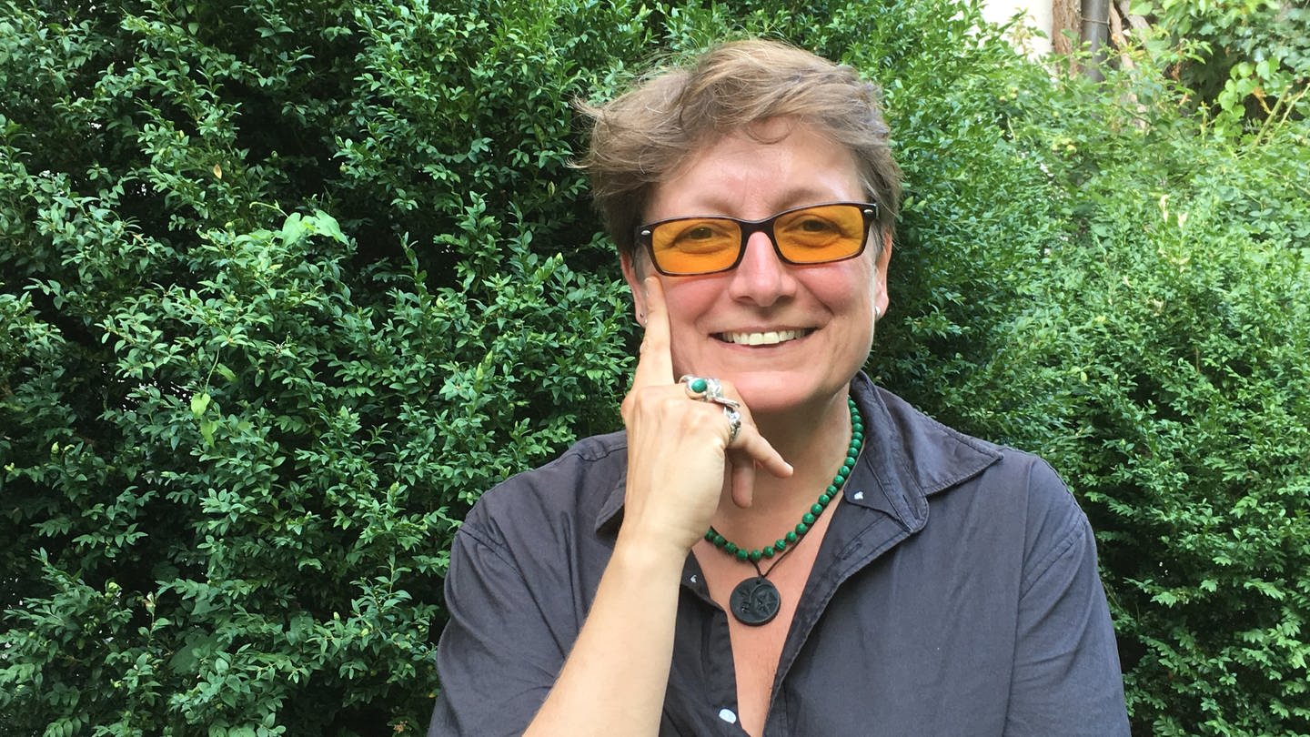 Frau mit gelb getönter Brille blickt in die Kamera, im Hintergrund grünes Gebüsch (Foto: SWR, Esther Leuffen)