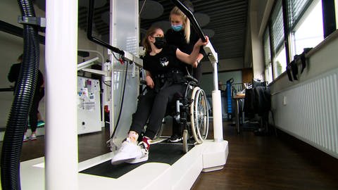 Frau im Rollstuhl wird von Therapeutin auf ein Therapiegerät geschoben. (Foto: SWR)