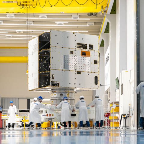 Ein Satellit steht in einer Halle, mehrere Menschen in weißer Arbeitskleidung stehen ebenfalls in der Halle. (Foto: Pressestelle, Aisbus Defence and Space)
