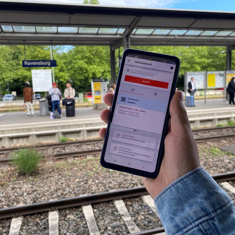 Seit Mittwoch gilt das 9-Euro-Ticket. Auf einem Handy ist die digitale Version zu sehen. Im Hintergrund der Bahnhof Ravensburg.