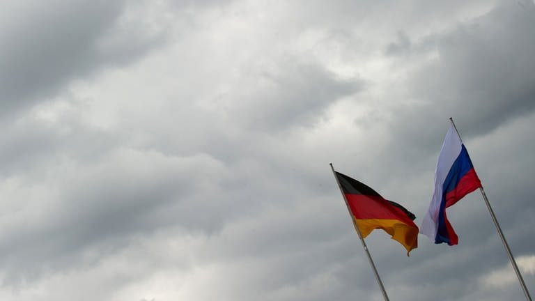 Eine russische und eine deutsche Flagge wehen im Wind vor wolkenbedecktem Himmel. 
