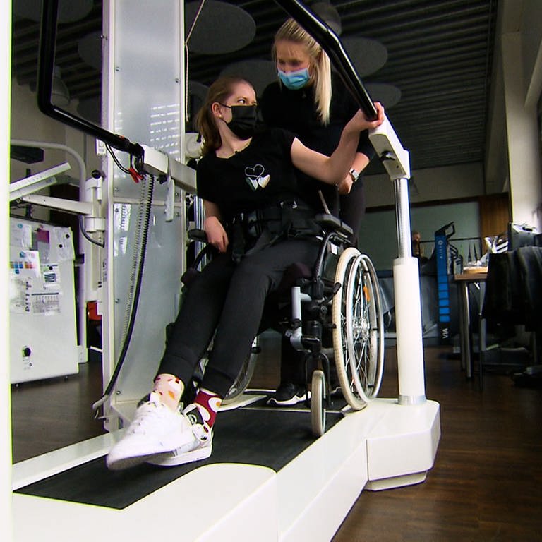 Frau im Rollstuhl wird von Therapeutin auf ein Therapiegerät geschoben. (Foto: SWR)