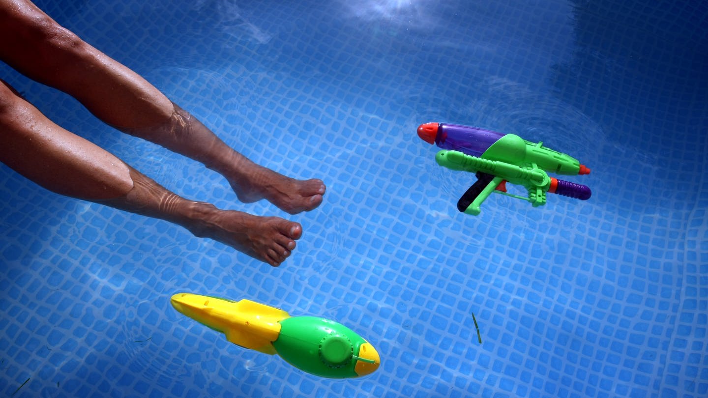 Füße baumeln im Wasser eines Swimming-Pools, daneben schwimmen zwei Wasser-Pistolen. (Foto: dpa Bildfunk, picture alliance / dpa | Karl-Josef Hildenbrand)